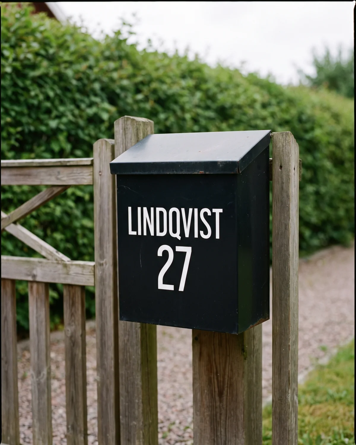 Svart brevlåda vid en grind med vitt namn och husnummer i skuren vinyl.